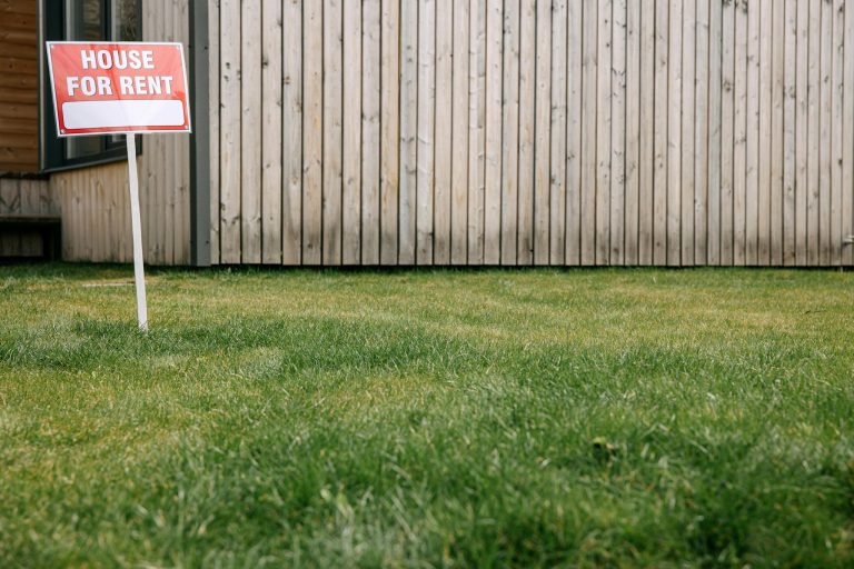 Close-up of 'House for Rent' sign on green lawn in front of a wooden house exterior.